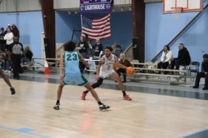 A basketball player dribbles the ball while closely guarded by an opponent, with spectators watching in the background