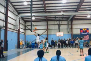 A player in a white jersey leaps to dunk a basketball while surrounded by opponents, with spectators watching an intense game