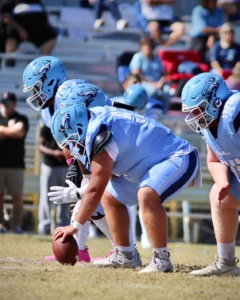 A trio of football players in light blue uniforms prepare for a play, with one player centered over the football on the field.